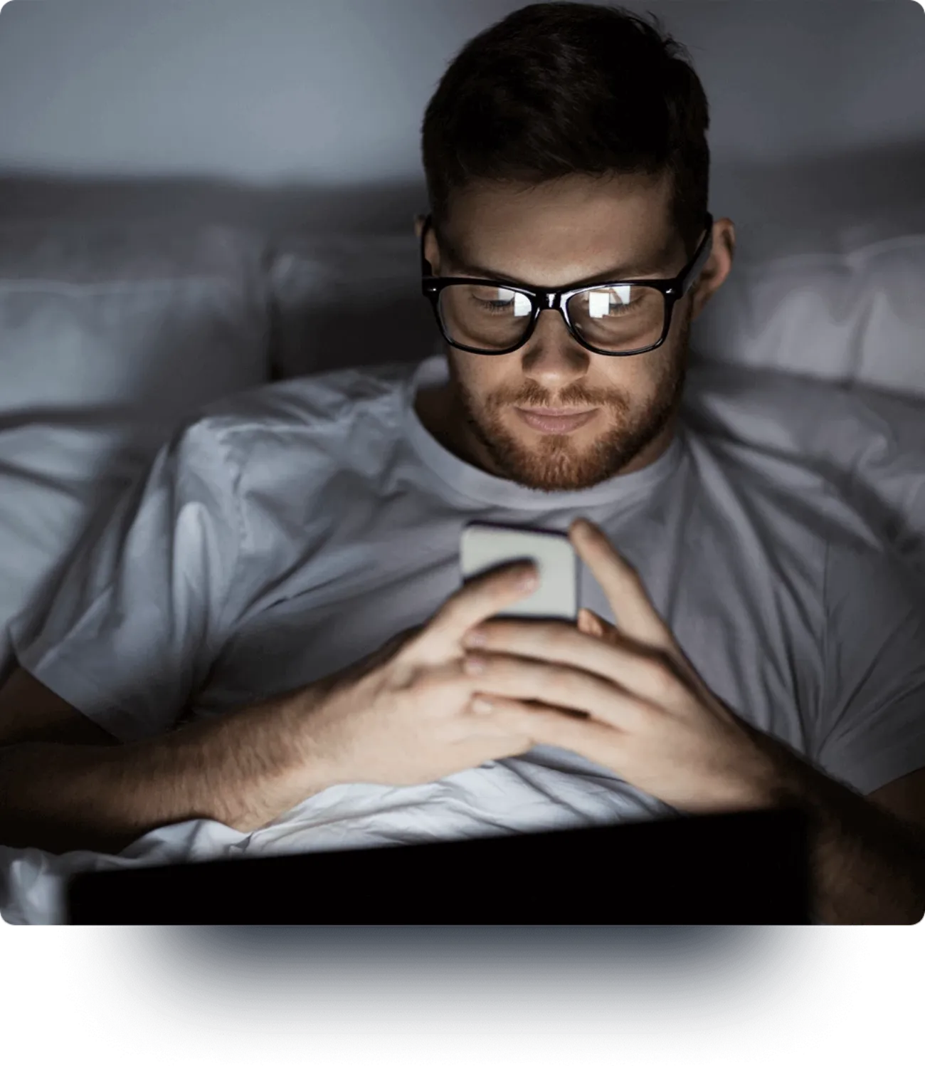 A man in bed, looking at his illuminated phone.