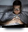 A man in bed, looks at his illuminated phone.