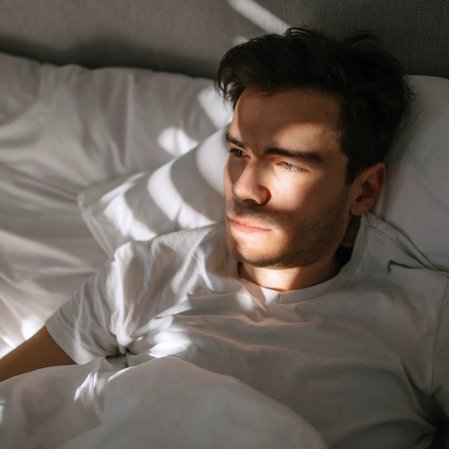 A young man lies in bed wearing a white t-shirt, partially illuminated by sunlight streaming through a window. 